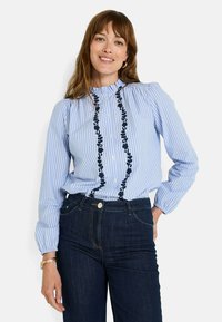 Camisa de rayas azul y blanca con cuello volado, decorada con bordado floral oscuro en el frente, combinada con jeans de mezclilla oscura.