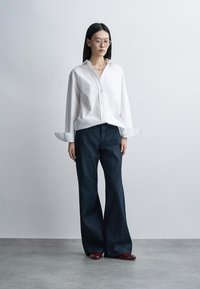 Femme portant une chemise blanche oversize, un jean large foncé, des chaussures rouges et des lunettes rondes, debout contre un mur gris clair uni.