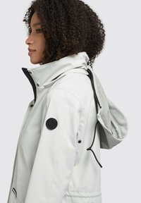 Light gray jacket with a high collar, snap buttons, and a black logo patch. Features a mesh-lined hood and adjustable drawstring.