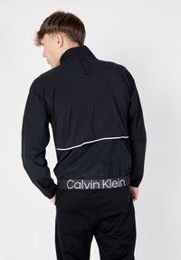 Giacca nera con zip e colletto alto, con dettagli bianchi lungo la vita. Include una banda con logo in vita, vista dal retro.