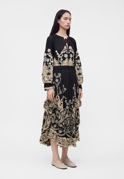Woman standing wearing a black dress with beige floral embroidery and beige flat shoes with ankle ties on a plain light background.