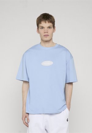 Fiatal férfi áll egyenes testtartással, világoskék túlságosan bő pólót és fehér nadrágot viselve, egyszerű világos háttér előtt.