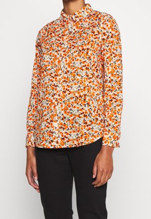 Kvinde iført en langærmet skjorte med knapper, der har et lille blomstermønster i orange, brun og beige, samt sorte bukser.