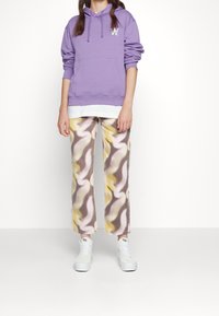 Fioletowa bluza z kapturem z kieszenią z przodu, nałożona na białą koszulę. Spodnie mają falisty wzór w żółtym, różowym i brązowym kolorze. Białe sneakersy.
