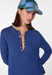 Blauwe gebreide trui met een diepe V-hals, voorzien van gouden knopen aan de voorkant. Het heeft lange mouwen en een relaxte pasvorm. Een blauwe geruite pet maakt het geheel af.