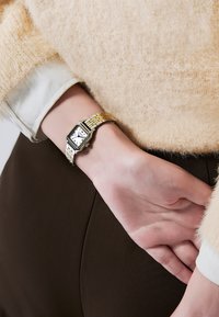 Montre rectangulaire dorée avec un cadran blanc, des chiffres romains noirs, et un bracelet en métal tissé, portée sur un poignet avec un léger pull.