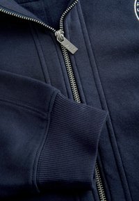 Granatowa bluza z prążkowanym mankietem i kołnierzem, z zamkiem błyskawicznym z metalowym, srebrnym suwakiem oraz teksturą czarnej tkaniny. Na piersi widoczne logo marki.