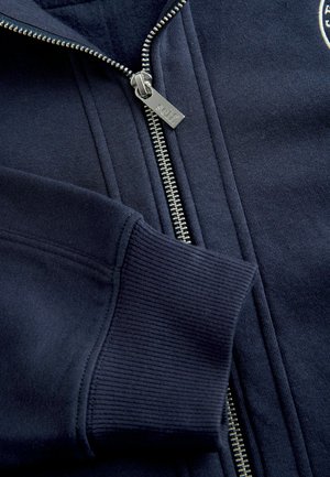 Marineblå sweatshirt med ribbet manchet og krave, med en sølvfarvet metal-lynlås og sort stoftekstur. Brandlogo synligt på brystet.