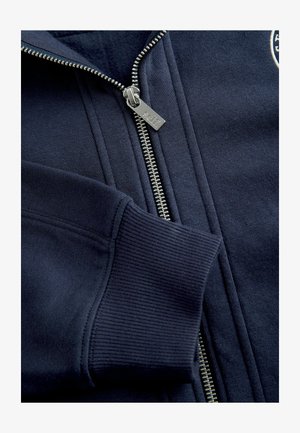 Granatowa bluza z prążkowanym mankietem i kołnierzem, z zamkiem błyskawicznym z metalowym, srebrnym suwakiem oraz teksturą czarnej tkaniny. Na piersi widoczne logo marki.