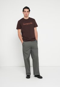 Smeđa pamučna majica s natpisom "CALVIN KLEIN" u narančastoj boji, uparena s širokim sivim hlačama i crnim cipelama. Jednostavan dizajn, ležeran izgled.