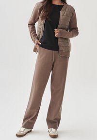 Giacca a zip marrone trapuntata su una maglia nera, abbinata a pantaloni larghi marroni e sneakers beige. Texture liscia con polsini a coste.