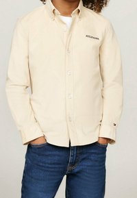 Dziecko z kręconymi włosami, ubrany w beżową koszulę z guzikami z napisem "HILFIGER" oraz niebieskie dżinsy, z rękami w kieszeniach, stojące na jednolitym tle.