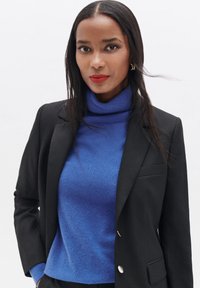 Blazer negro sobre un suéter de cuello alto azul. El blazer cuenta con un solo botón y solapas elegantes. Tejido suave con un ligero brillo.