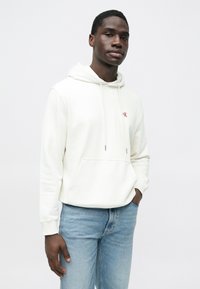 Sudadera blanca con capucha que cuenta con un bolsillo delantero y un logo rojo "CK". Combinada con vaqueros azul claro, ofrece un estilo casual.