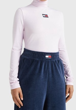 Person trägt einen hellrosa Rollkragenpullover und marineblaue Velourhosen, beide mit Tommy Jeans Logo-Patches auf der Brust und am Bund.