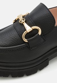 Sorte lærloafers med en teksturert overflate, med detaljer i gulltone i form av sirkulært hardware og en tykk svart gummisåle.