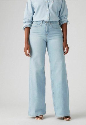 Personne portant un jean large bleu clair et une chemise boutonnée bleu clair assortie avec manches retroussées, debout les mains détendues.