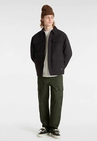 Chaqueta negra abotonada con dos bolsillos en el pecho, usada sobre una camiseta de color claro, combinada con pantalones cargo verdes y zapatos negros.