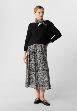 Schwarzer Strickpullover über einem grauen Leopardenmuster-Rock, mit einem am Kragen gebundenen Schal. Schwarze Ballerinas runden das Outfit ab.