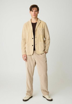 Jonge man staand, draagt een beige ribfluwelen pak met zwarte knopen, een zwart overhemd eronder en witte sneakers met donkere zolen.