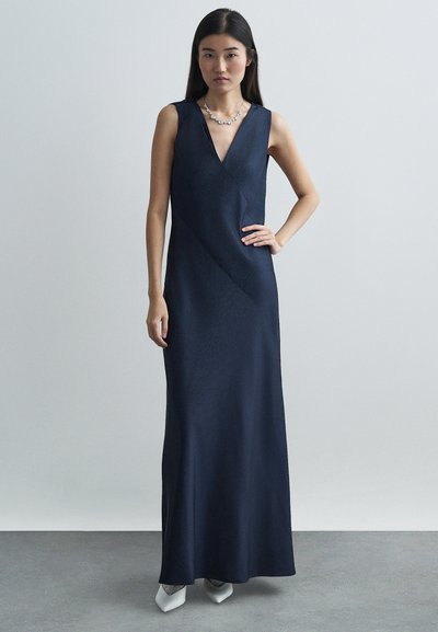 Robe sans manches bleu marine avec un décolleté en V, présentant une texture lisse et soyeuse ainsi que des coutures diagonales, associée à des escarpins blancs pointus et un collier.