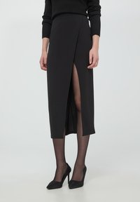 Gonna nera a matita con un'alta scollatura, texture liscia, lunghezza a metà polpaccio, abbinata a tacchi alti neri e lucidi.