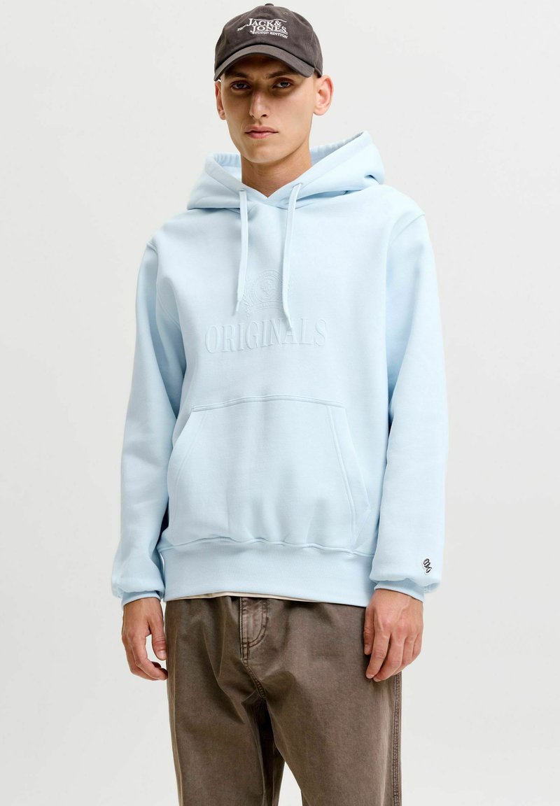 Jeune homme portant un sweat à capuche bleu clair avec « Originals » en relief sur le devant, pantalon marron et casquette sombre à l’intérieur sur un fond uni.
