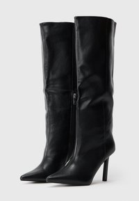Un par de botas de cuero negro hasta la rodilla con punteras puntiagudas y tacones de aguja, que cuentan con cremalleras en el lado interno.