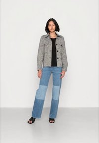 Camisa tipo jaqueta em padrão pied-de-poule cinza com botões pretos, combinada com calças de ganga largas em azul claro e blocos de cor, e sandálias pretas de tiras.