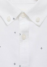 Bílé polo tričko s dvouknoflíkovým zapínáním, pokryté malými námořnickými puntíky na látce. Texturovaný bavlněný materiál, klasický design límce.