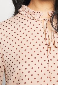 Blouse beige à pois marron, avec un col montant froncé et un ruban à l'encolure, portée par une personne aux cheveux foncés.