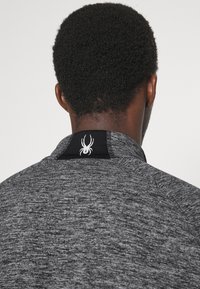 T-shirt de sport gris avec une finition texturée, présentant un col noir et un logo araignée blanc au dos. Design simple, sans motifs.