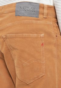 Kordurojske traperice v svetlo rjavi barvi z značilnimi podrobnostmi na zadnjem žepu. Imajo embosiran logotip in rdeče šivane poudarke na žepu.