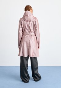 Vodaodolný růžový trench coat s kapucí, páskem v pase a dlouhým střihem, v kombinaci s volně padnoucími černými kalhotami.