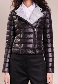 Femme portant une veste matelassée noire brillante avec une fermeture éclair asymétrique, sur une chemise blanche à col et un pantalon noir.