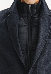 Czarny, wełniany płaszcz w ciemnoszarym odcieniu z wysokim kołnierzem, z warstwą czarnego, pikowanego wnętrza. Posiada centralny zamek błyskawiczny i teksturowany materiał.