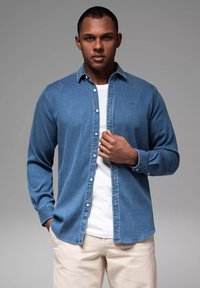 Denimhemd in mittlerem Waschblau, mit Knopfleiste, langen Ärmeln, weicher Textur, über einem weißen T-Shirt getragen, mit lässiger Passform und minimeller Markenkennzeichnung.