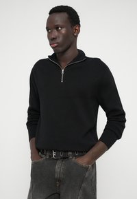 Jaunas vyras, vilkintis juodą užtrauktuką ir išblukusias juodas džinsus, stovi su rankomis kišenėse, žiūrėdamas į šoną prieš paprastą baltą foną.