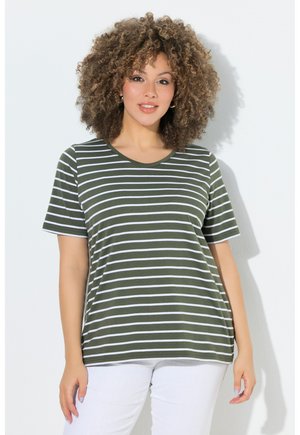 Frau mit lockigem Haar, die ein olivgrün-weiß gestreiftes Kurzarmshirt und weiße Hose trägt, steht vor einem einfarbigen hellen Hintergrund.