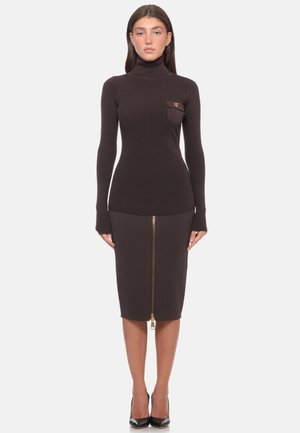 Donna che indossa un maglione aderente a collo alto marrone scuro con taschino sul petto e una gonna a matita abbinata con una zip dorata frontale e tacchi neri.