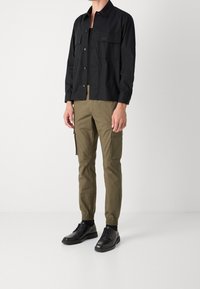 Chemise noire à boutons avec deux poches poitrine, associée à un pantalon cargo vert olive avec poches zippées et chaussures noires à semelles épaisses.