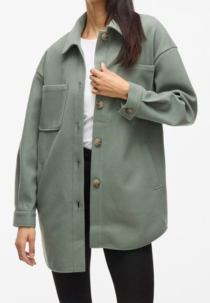 Femme portant une veste verte oversize boutonnée avec une grande poche poitrine, chemise blanche et pantalon noir, tenant la veste près du col.