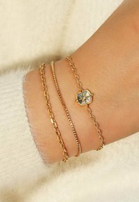 Tre bracciali d'oro su un polso: una catena sottile, un bracciale a maglie piatte e uno con un charm esagonale che presenta un motivo a stella. Superficie texturizzata visibile.