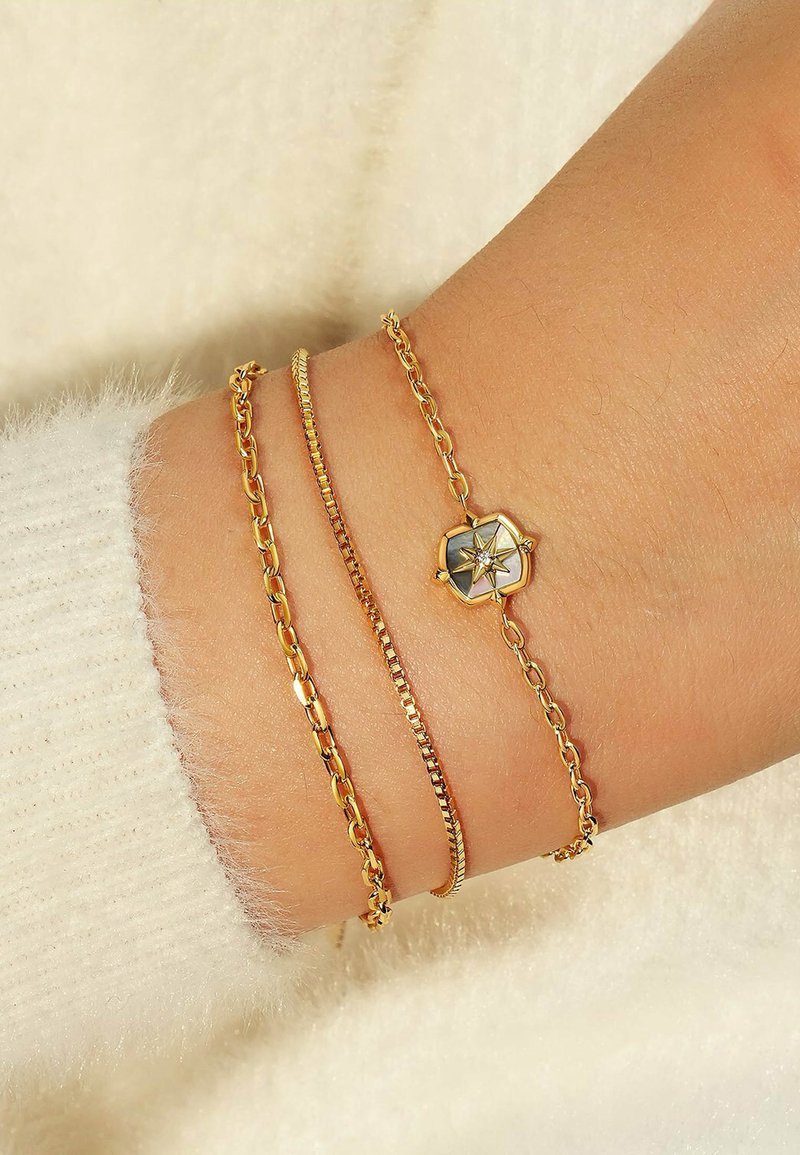 Tre bracciali d'oro su un polso: una catena sottile, un bracciale a maglie piatte e uno con un charm esagonale che presenta un motivo a stella. Superficie texturizzata visibile.