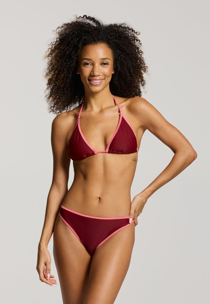 Donna con capelli ricci sorridente, che indossa un bikini color amaranto con bordi rosa, in piedi davanti a uno sfondo chiaro e uniforme.
