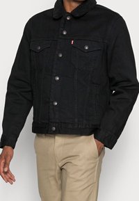 Schwarze Jeansjacke mit einem weichen, schwarzen Kunstfellkragen, ausgestattet mit Druckknöpfen, zwei Brusttaschen und Seitentaschen. Getragen mit beigen Hosen.