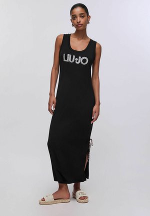 Donna che indossa un abito lungo nero senza maniche con logo "LIU JO", spacco laterale e sandali beige slip-on con suola platform.