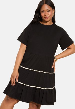 Zwart jurk met korte mouwen, met een gelaagd ontwerp. Opmerkelijke details zijn een decoratieve crèmekleurige bies rond de taille.