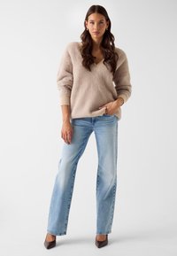 Beige, pluizige V-hals trui met geribde boorden, gecombineerd met blauwe jeans met rechte pijpen. Draagt daarbij grijze pumps met spitse neus. Simpele, moderne uitstraling.