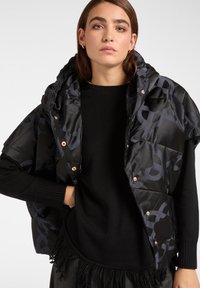 Kobieta nosząca czarną kamizelkę pikowaną w wzory na czarnym swetrze z długim rękawem i frędzlami na dole, stojąca na tle jasnego, jednolitego tła.